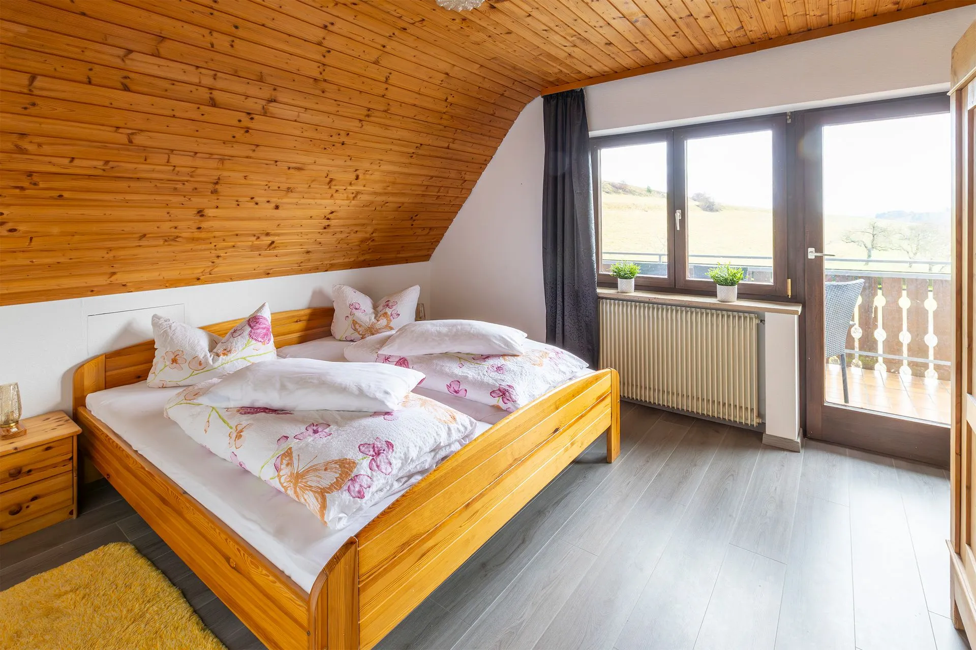 Schlafzimmer mit einem Doppelbett aus Holz, zwei Kopfkissen mit buntem Muster, Holzdecke und Fenster mit Blick auf die Landschaft.