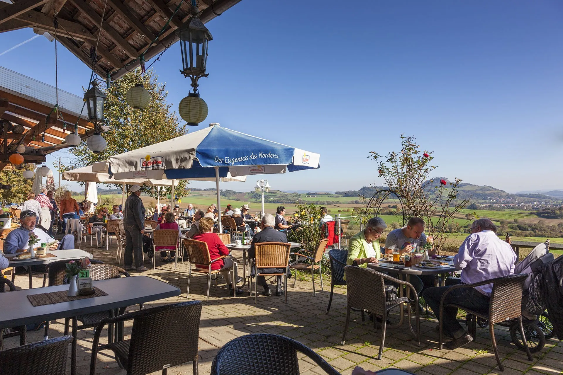 Terrasse eines Restaurants mit vielen Gästen, Tischen und Stühlen. Ein großer Sonnenschirm mit der Aufschrift 'Der Frugale das Norden' steht im Hintergrund. Landschaft mit Bäumen und Hügeln ist sichtbar.