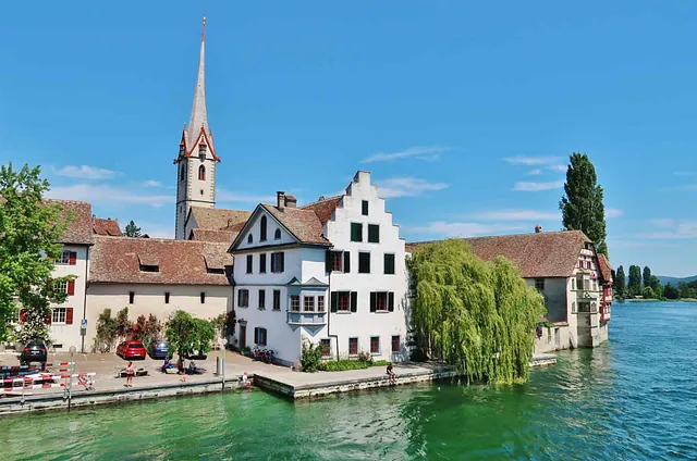 Blick auf die Stadt Stein am Rhein in der Schweiz, mit historischen Gebäuden, einem Kirchturm und Bäumen am Ufer des Rheins. Sichtbare Autos auf der Straße.