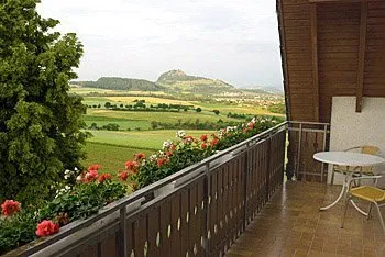 Balkon mit Holzgeländer, blühenden Pflanzen und einem Tisch mit zwei Stühlen. Im Hintergrund eine grüne Landschaft mit Hügeln.