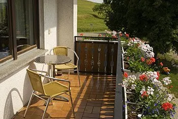 Ein Balkon mit einem runden Tisch und zwei Stühlen aus Metall mit gelben Sitzkissen. Der Boden ist gefliest, und es gibt Blumen in Kästen entlang der Brüstung.