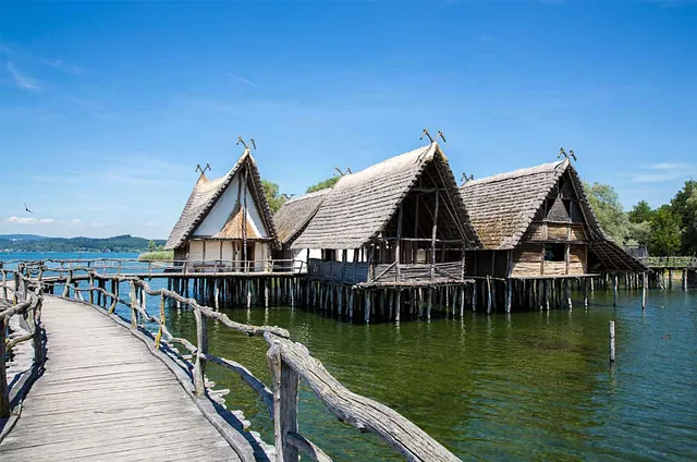 Drei historische Holzhäuser auf Stelzen im Wasser, umgeben von Bäumen und einem Holzsteg. Sichtbare Beschriftung: 'PFAHLBAUMUSEUM Unteruhldingen'.