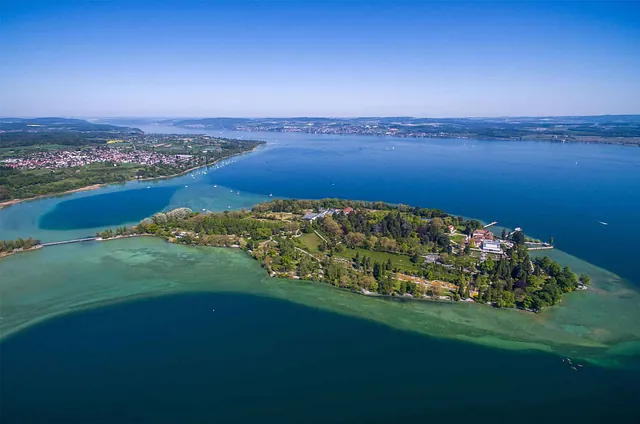 Luftaufnahme der Insel Mainau im Bodensee, umgeben von Wasser und grünen Uferbereichen. Sichtbare Gebäude und Bäume auf der Insel.