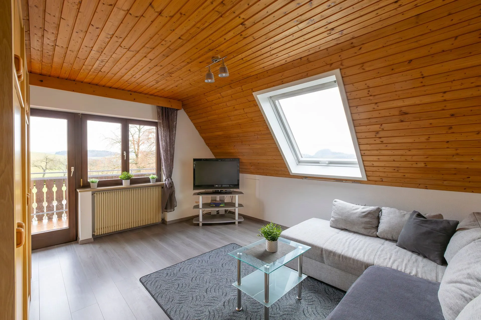 Wohnzimmer mit schrägem Holzdecke, grauem Sofa, Glastisch und Fernseher. Fenster mit Blick auf die Landschaft, Pflanzen auf dem Tisch.