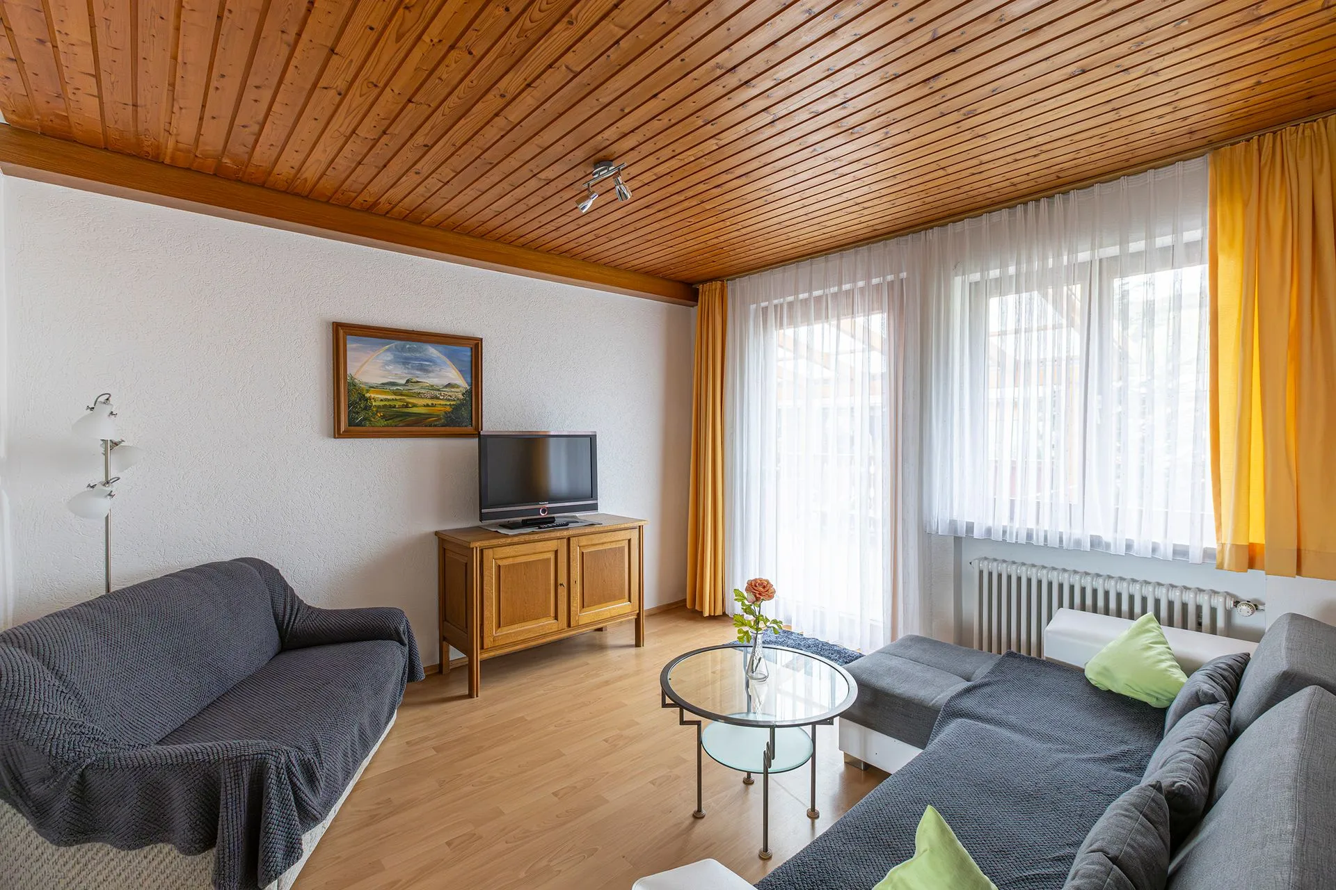Wohnzimmer mit Holzdecke, zwei Sofas, einem Couchtisch und einem Fernseher. Ein Gemälde hängt an der Wand. Vor einem Fenster hängen gelbe Vorhänge.