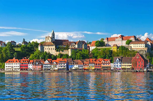 Panoramaansicht von Meersburg am Bodensee, mit bunten Häusern am Ufer und einer historischen Burg auf einem Hügel im Hintergrund. Sichtbare Wolken am blauen Himmel.