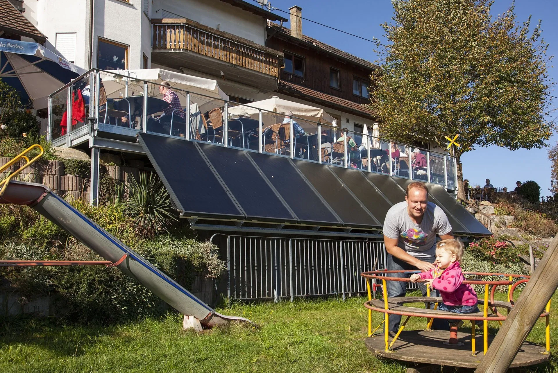 Ein Kind spielt auf einem Karussell, während ein Erwachsener daneben steht. Im Hintergrund sind Sonnenschirme und eine Terrasse eines Gebäudes sichtbar. Solarzellen sind auf dem Dach angebracht.