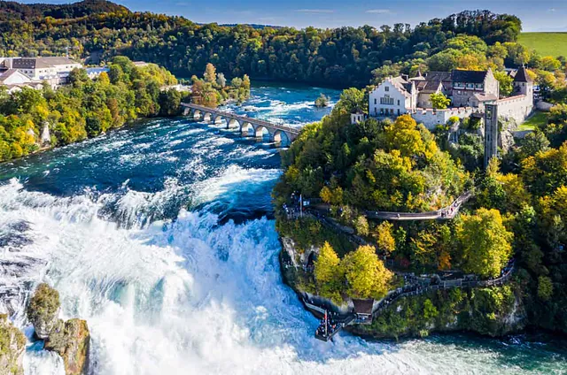Luftaufnahme des Rheinfalls in Schaffhausen, Schweiz, mit Wasserfällen, Brücke und umgebender Vegetation. Sichtbare Beschriftung: 'RHEINFALL SCHAFFHAUSEN, Schweiz'.