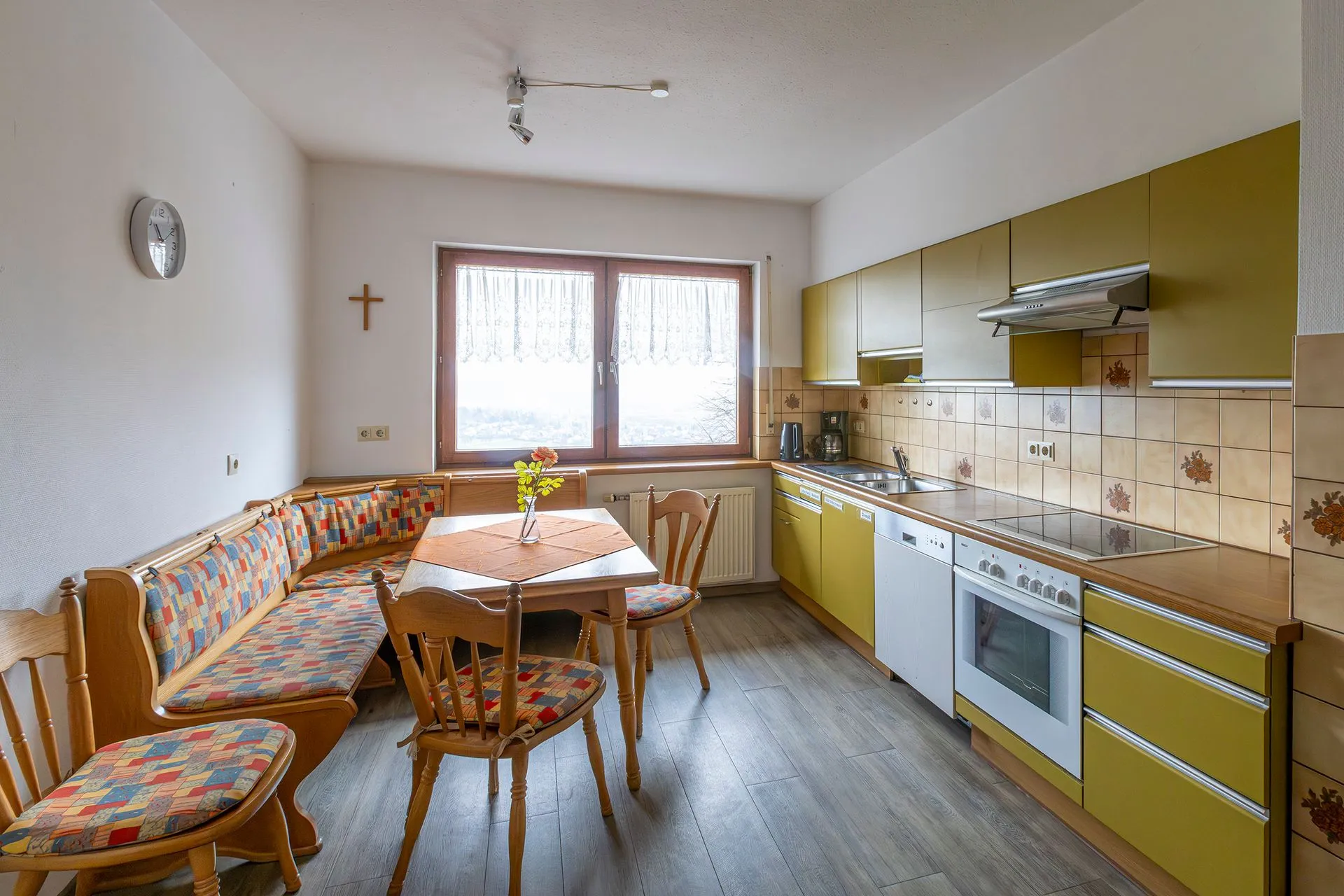 Küche mit gelben Schränken, einem weißen Herd, einem Tisch mit orangefarbener Tischdecke und einer Eckbank mit buntem Muster. Fenster mit Vorhängen.