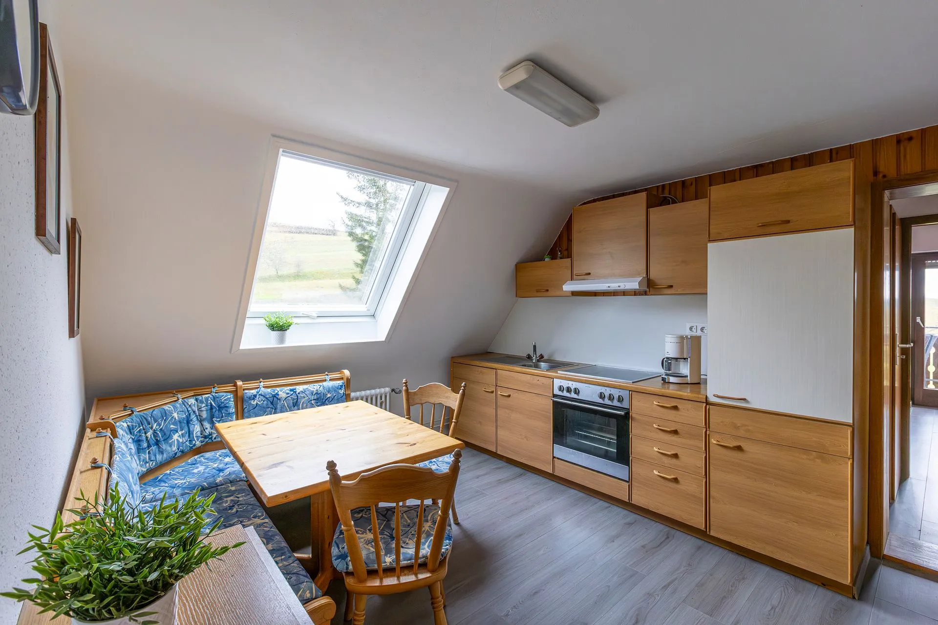 Küche mit Holzschränken und einem Tisch mit vier Stühlen. Ein Fenster mit Blick auf Bäume. Eine grüne Pflanze auf dem Tisch.