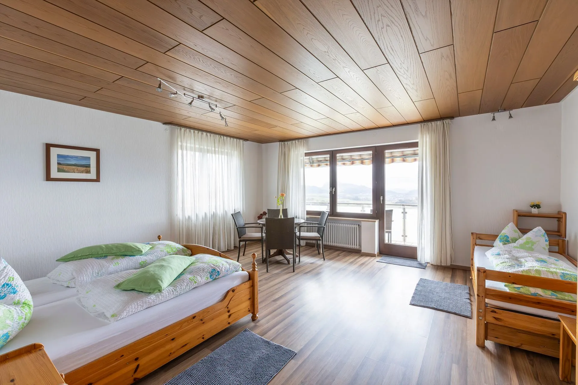 Zimmer mit zwei Einzelbetten aus Holz, grüne Bettwäsche, Tisch mit Stühlen, große Fenster mit Aussicht, Holzboden.