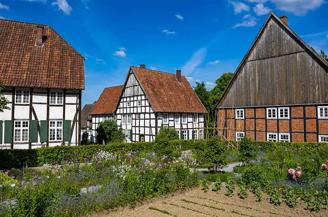 Drei historische Fachwerkhäuser mit roten Ziegeldächern, umgeben von einem Garten mit bunten Blumen. Unten steht 'FREILICHTMUSEUM Neuhausen'.