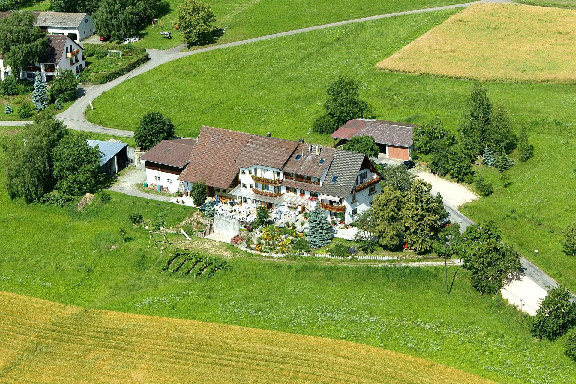 Luftaufnahme eines großen, zweigeschossigen Hauses mit einem Garten, umgeben von Wiesen und Bäumen. Ein Weg führt zum Haus.