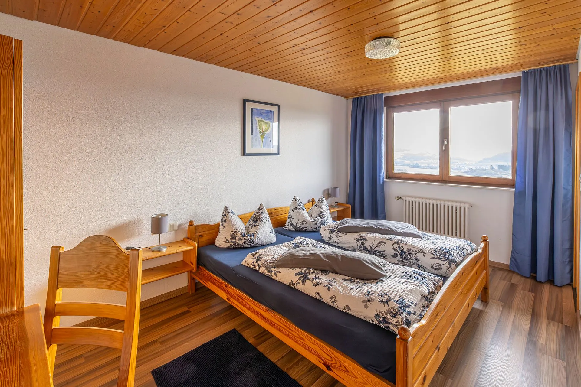 Ein Schlafzimmer mit zwei Einzelbetten, blauen Bettdecken und Kissen. Ein Holzstuhl steht neben einem Nachttisch. Ein Fenster mit Blick auf die Landschaft.
