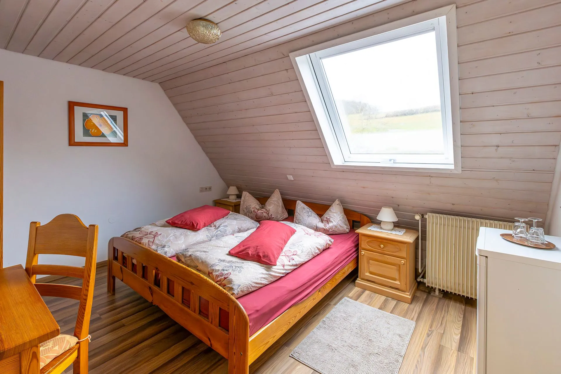 Dachzimmer mit einem Doppelbett, zwei Kissen und roten Bettdecken. Ein Holzstuhl und ein Tisch sind sichtbar. Ein Fenster mit Ausblick auf die Landschaft.