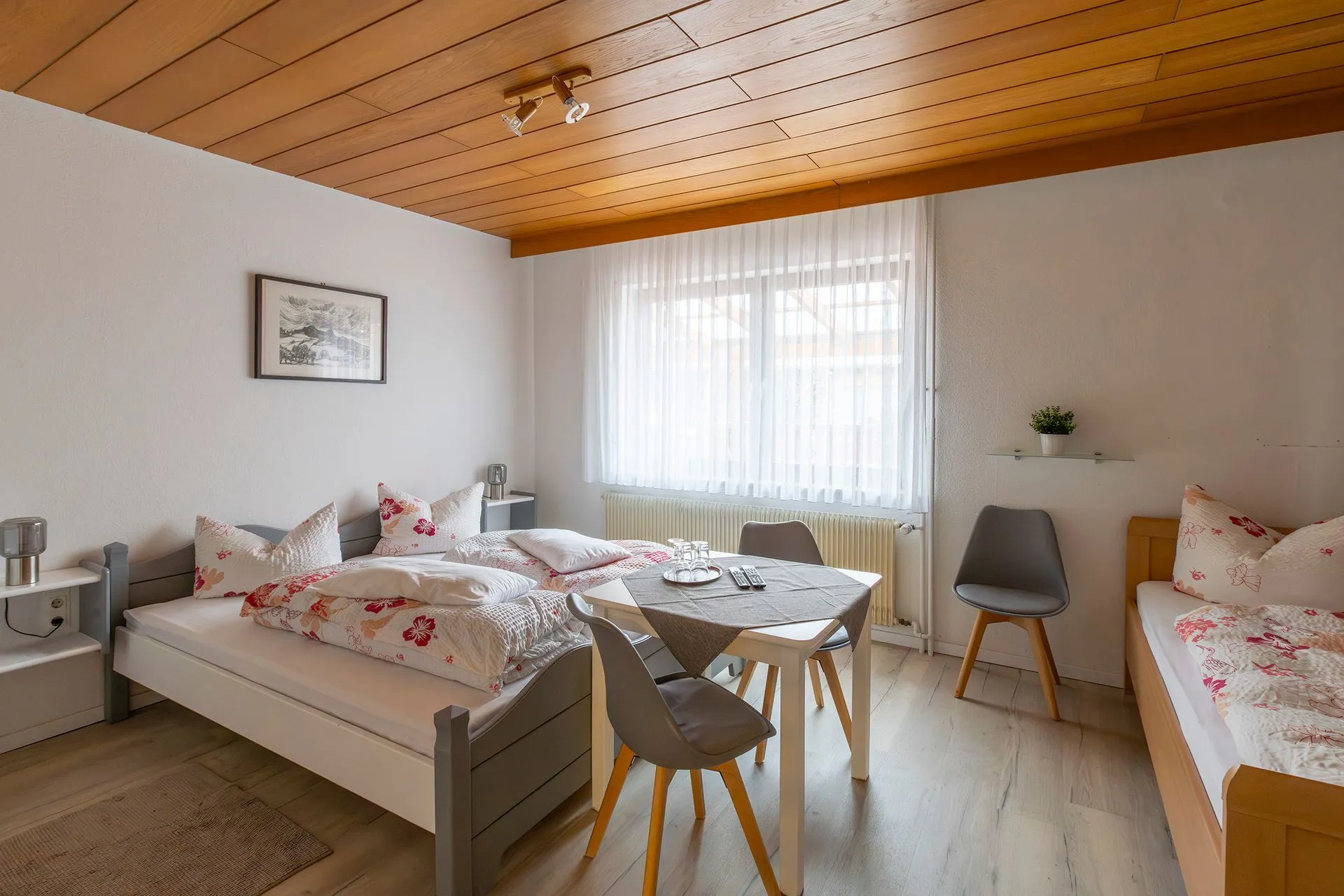 Helles Schlafzimmer mit zwei Betten, einem Tisch und Stühlen. Weiße Wände, Holzdecke, große Fenster mit Vorhängen. Dekorative Kissen mit Blumenmuster.