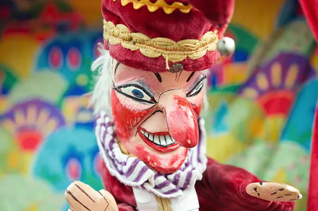 Puppe mit rotem Kostüm und braunen Händen, trägt einen Schal und hat eine bunte, gemusterte Umgebung im Hintergrund. Sichtbarer Text: 'FASCHNACHTSMUSEUM Langenstein'.