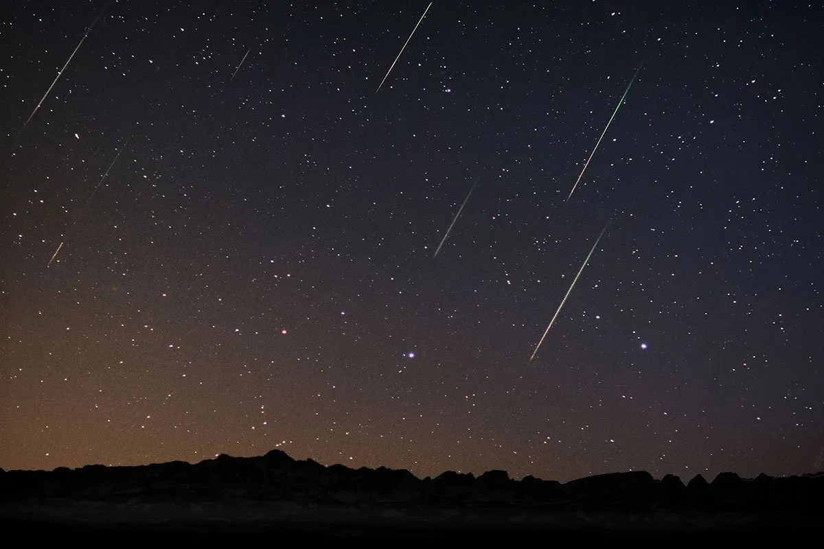 Nachthimmel mit vielen Sternschnuppen, die diagonal über den Himmel ziehen, und einer sternenklaren Kulisse mit Bergen im unteren Bereich.